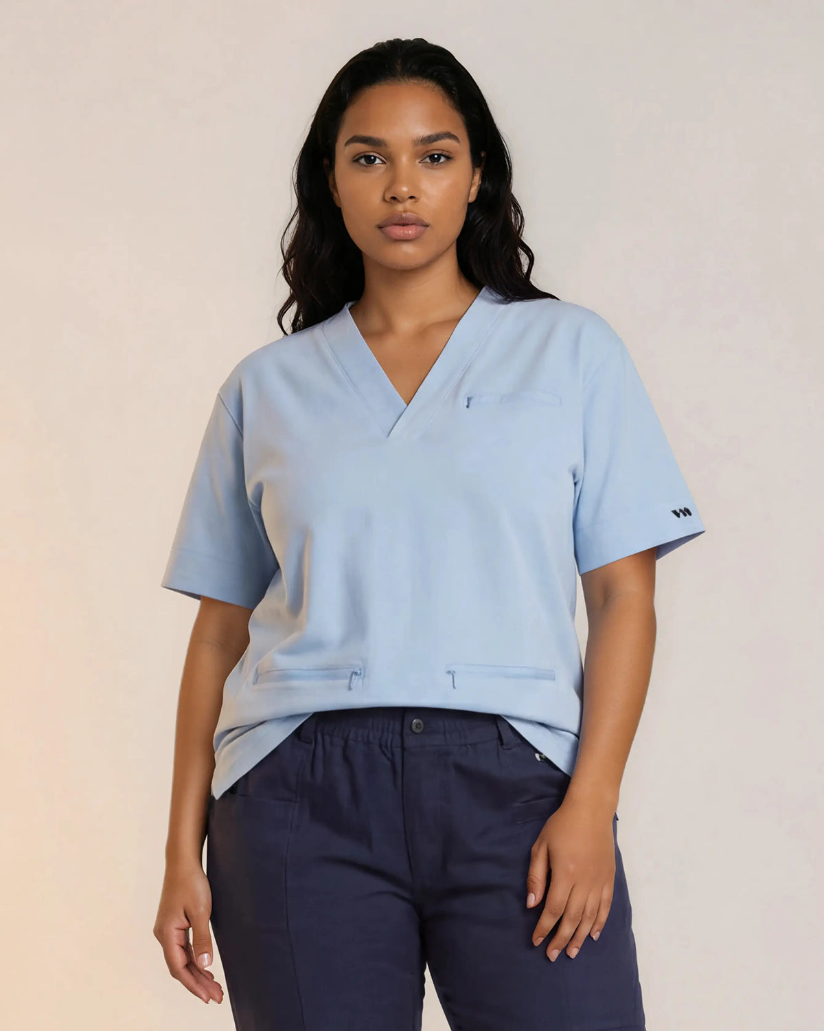 Femme medecin portant une tunique médicale bleu ciel SANARE à col en V, manches courtes, avec poche poitrine et deux poches zippées à la taille, associée à un pantalon médical bleu marine.